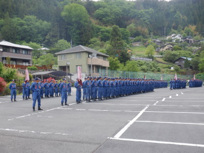 駐車場で薄い青色の制服を着た多数の人が姿勢よく立っている、団員訓練の写真
