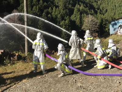 銀色の防火服を着た6名が、2名1組で放水している模擬火災訓練の様子の写真
