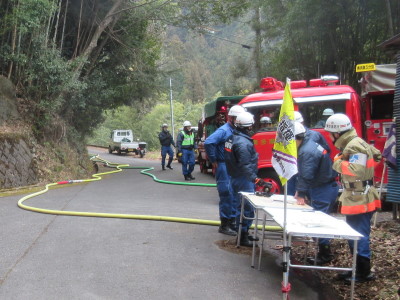 舗装された山道で、ポンプ車からホースを伸ばしたり、現状を確認している多数の模擬火災訓練の様子の写真